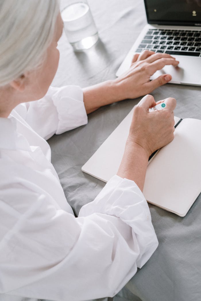 Elderly woman takes notes while using a laptop at home.