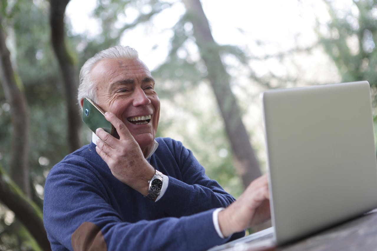 Happy senior man talking on phone and using laptop outdoors.